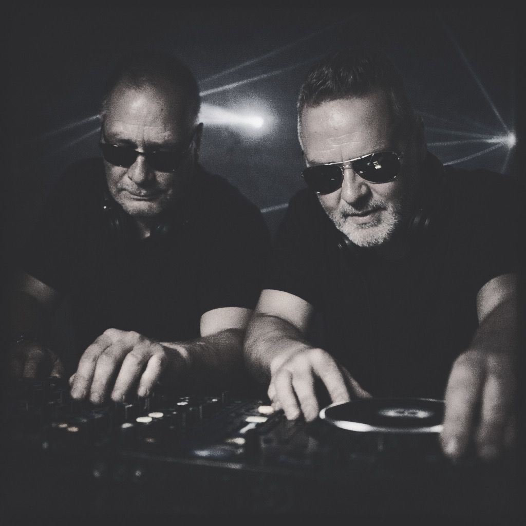 DJ bmp und electropapa mit Sonnenbrillen arbeiten konzentriert an einem Mischpult in einem dunklen Techno-Club. Kaltes Laserlicht durchschneidet den rauchigen Hintergrund. Das Bild wirkt körnig und kontrastreich wie ein analoger Blitz-Schnappschuss aus einem unterirdischen Bunker, mit klar erkennbaren Gesichtern und retrohafter Club-Atmosphäre.